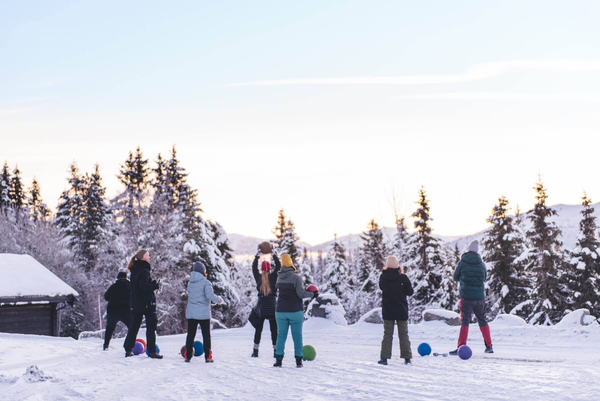 Barn og voksne leker i snøen med fargerike baller, omgitt av snødekte trær og fjell. Klar vinterhimmel.