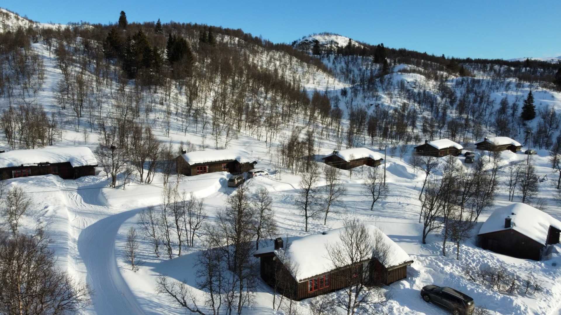 Snødekte hytter i et vinterlandskap med trær og fjell i bakgrunnen, blå himmel.