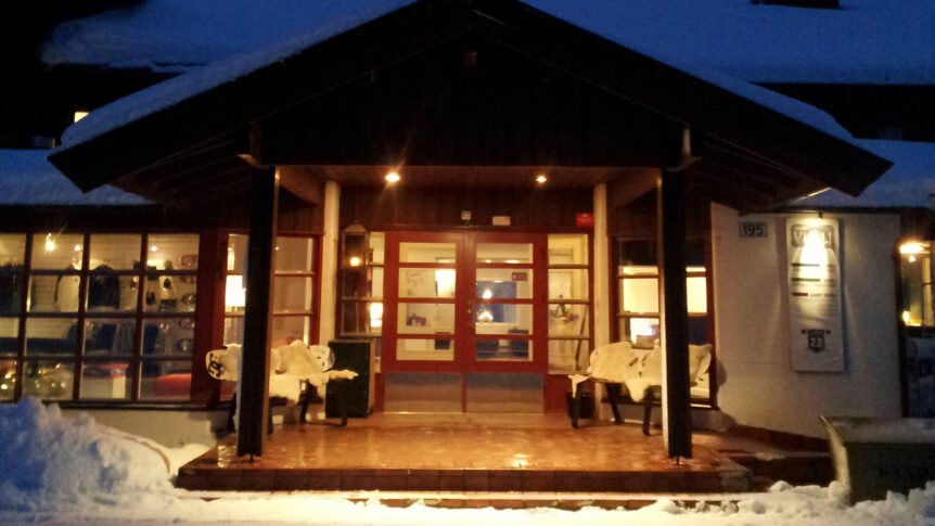Inngangspartiet til et snødekket trehus opplyst i skumringen, med tepper på benkene.