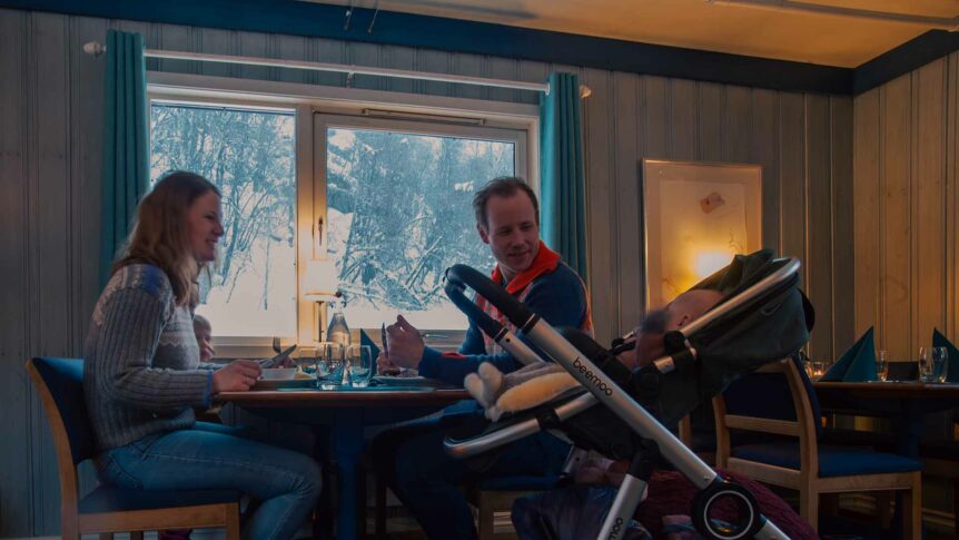 Familie på restaurant med snøutsikt, kvinne og mann ved bord, barnevogn med barn.