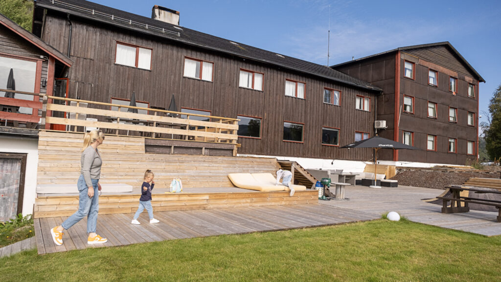 Kvinne og barn på treterrasse foran stort, trefarget bygg i solfylt vær.