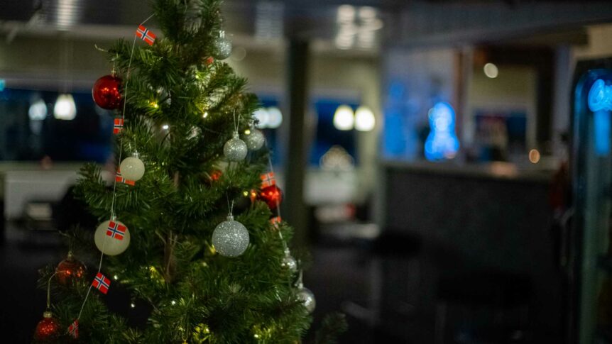Juletre dekorert med lys og røde ornamenter, pyntet med små norske flagg, i en koselig stue.