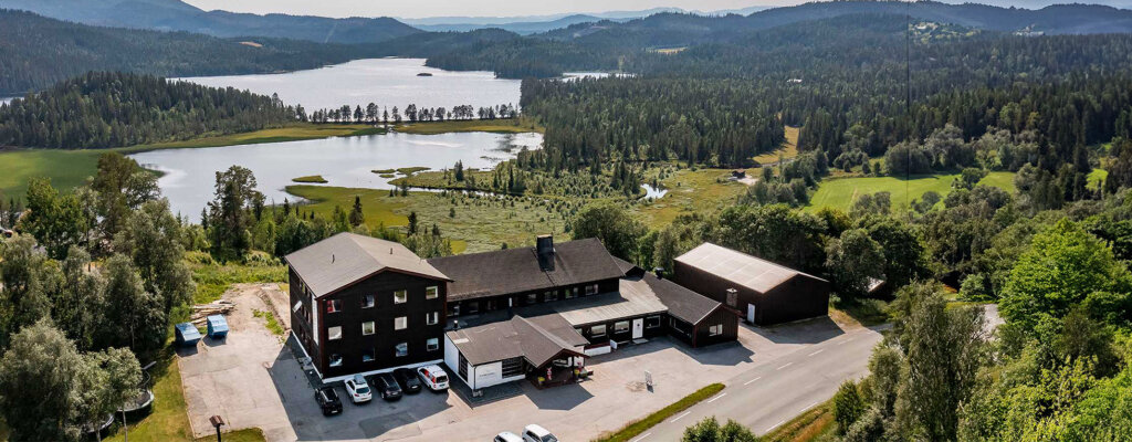 Panorama av hus ved innsjø omgitt av skog og fjell i et norsk landskap.