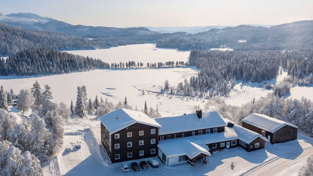 Vinterlandskap med snødekte trær og bygninger ved en islagt innsjø, omgitt av skogkledde fjell.
