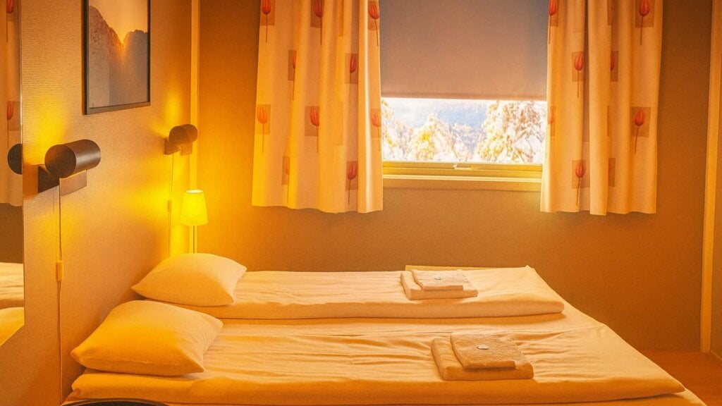 Koselig hotellrom med to enkeltsenger og varme belysning. Vindu med utsikt og gardiner med motiv av blomster.