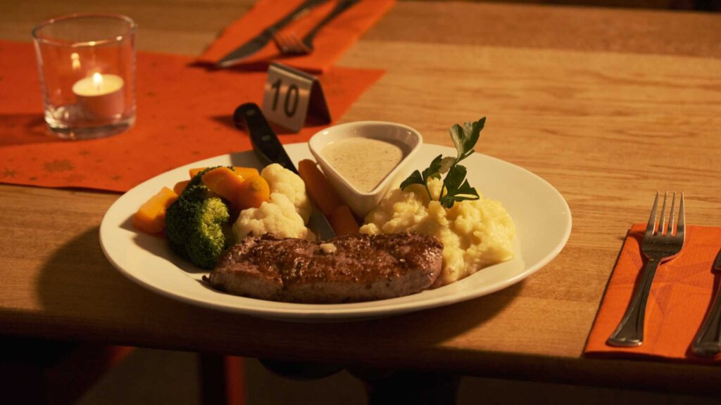 Tallrik med biff, potetmos, grønnsaker og saus, med tent lys i bakgrunnen.