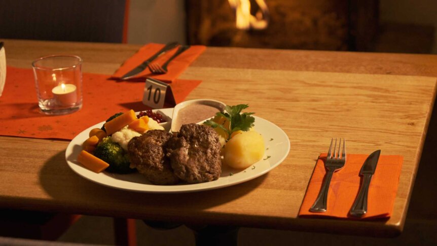 Hjemmelaget middag med karbonader, poteter, grønnsaker og saus på tallerken ved bord med orange serviett og lys.