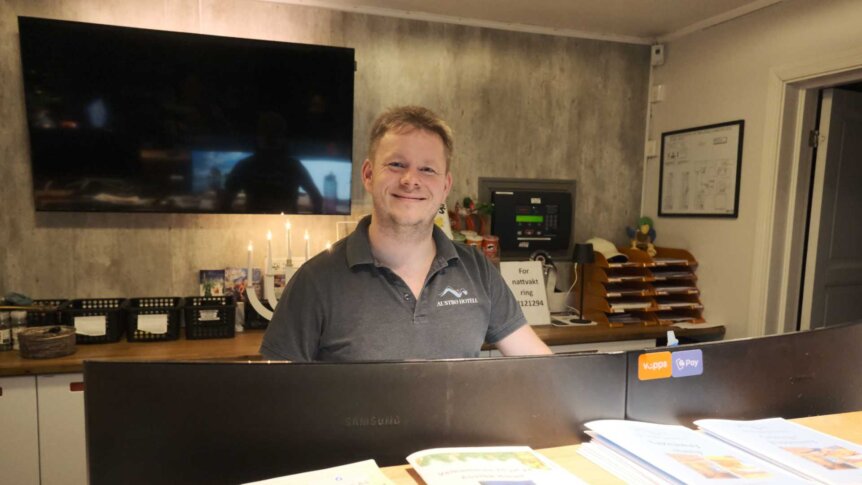 Resepsjonist smiler på Austbø Hotell, med brosjyrer og moderne interiør i bakgrunnen.