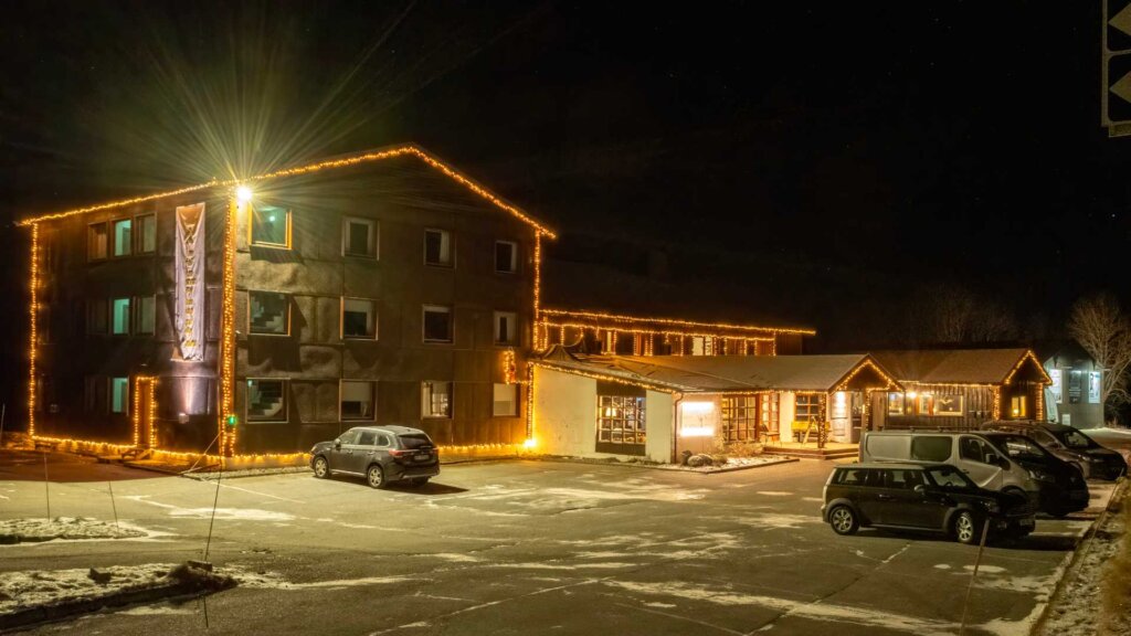 Nattbilde av et hotell dekorert med julelys, omgitt av parkerte biler og litt snø på bakken.