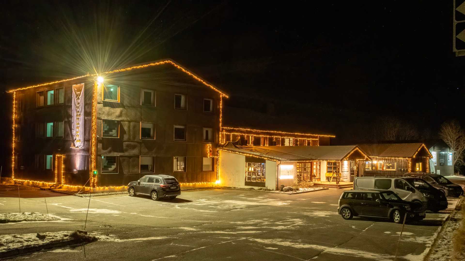 Nattbilde av et hotell dekorert med julelys, omgitt av parkerte biler og litt snø på bakken.
