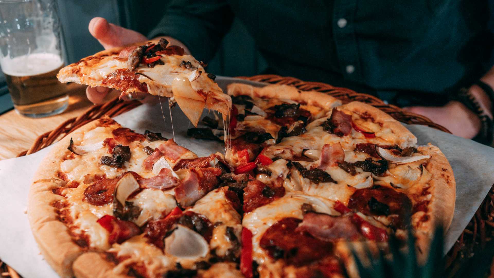 Person tar et stykke pizza med kjøtt og grønnsaker, glass øl i bakgrunnen.