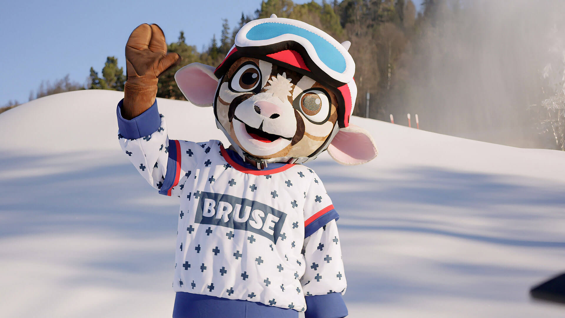 Maskot med vinterklær og skibriller på snødekt bakgrunn ved skogen. Morsom figur ser glad ut i solen.