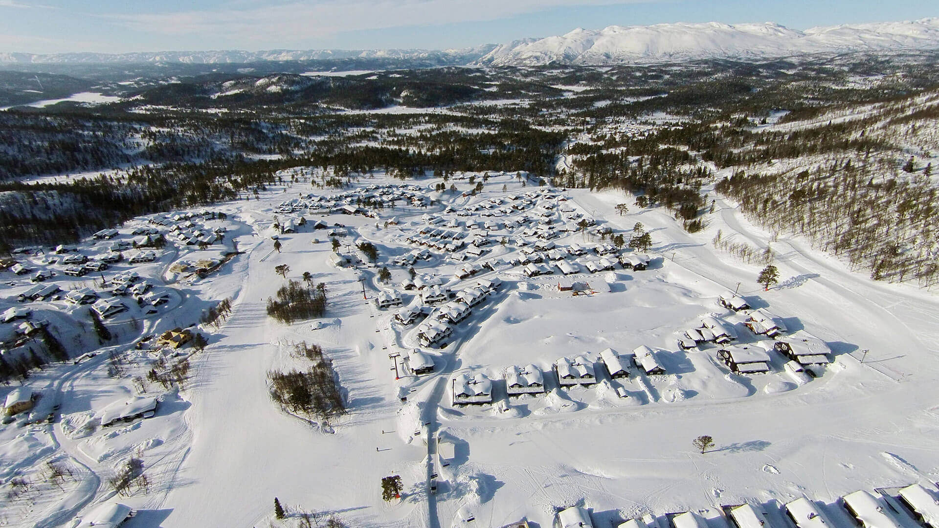 Snødekt norsk hyttefelt med fjelllandskap i bakgrunnen, tatt fra luften på en klar vinterdag.