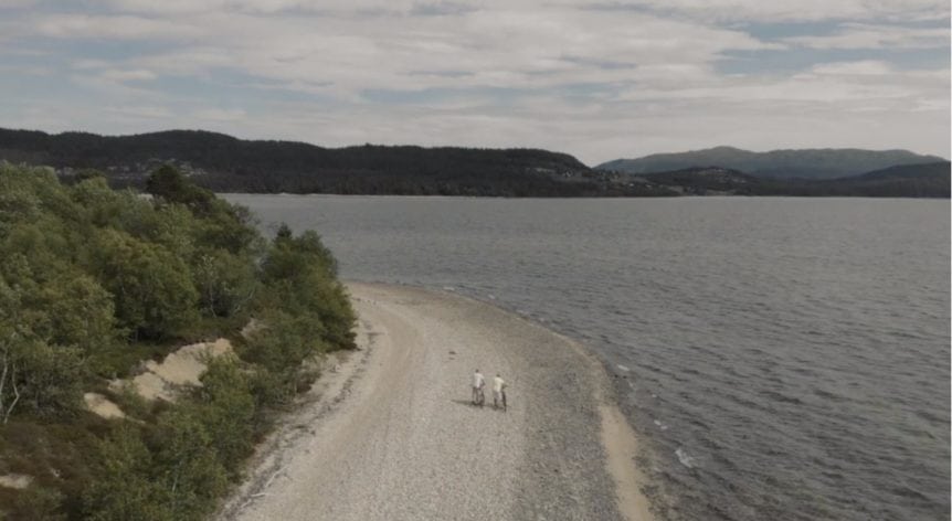 Strandpromenade ved innsjø omgitt av skog og fjell, under en overskyet himmel.