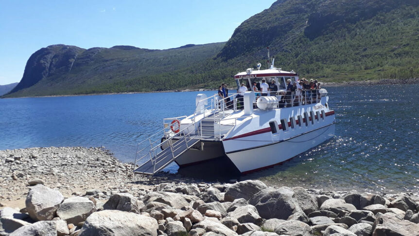 Katamaran med passasjerer ved en steinete strandlinje og naturskjønt fjellandskap i bakgrunnen, i solfylt vær.