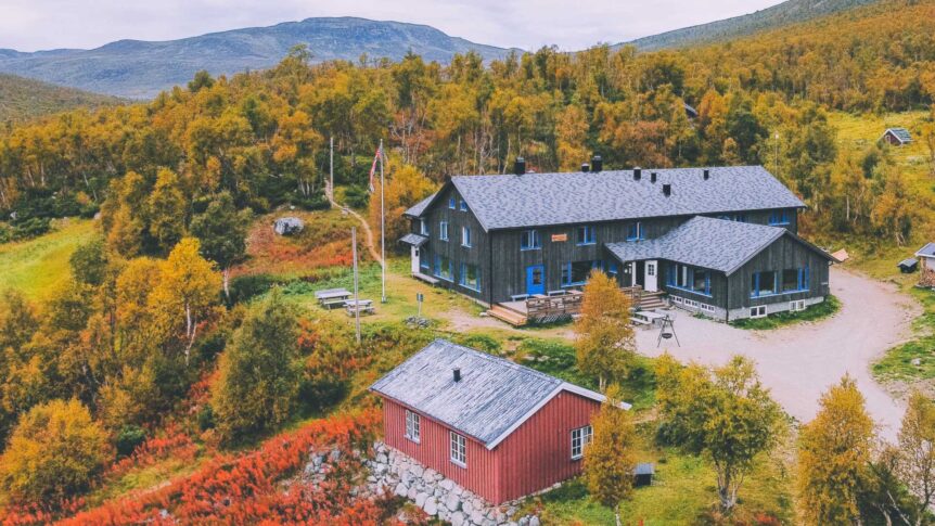 Fjellhytte omgitt av fargerike høsttrær i norsk natur, med fjell i bakgrunnen.
