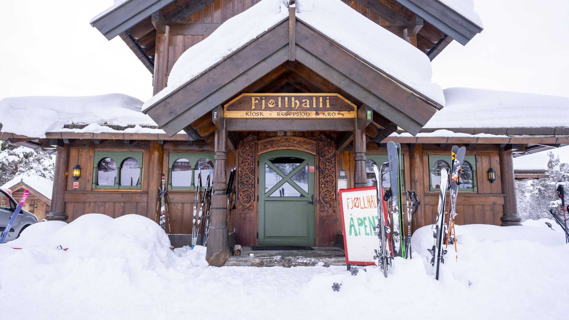 Tradisjonell fjellhytte i snødekt landskap med skistaver utenfor. Skilt viser Fjøllhalli - Åpen.