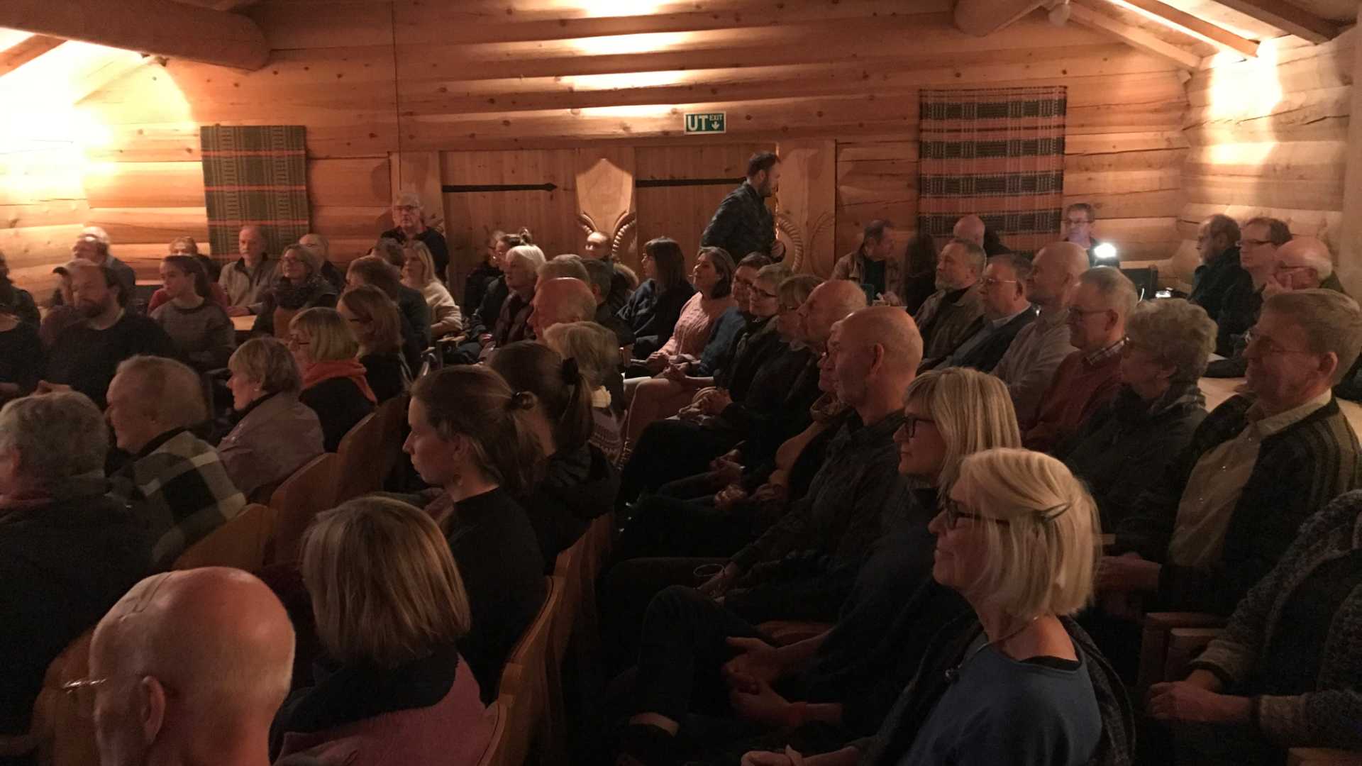 Mennesker i en trehytte deltar på et arrangement, ser fremover med interesse og oppmerksomhet.