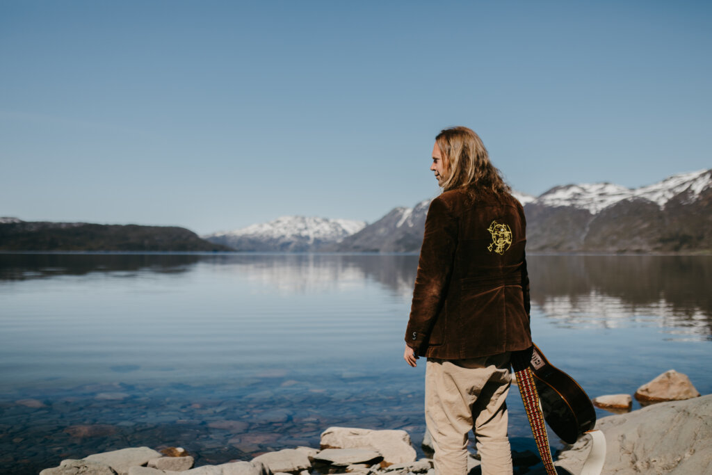 Mann med guitar står ved vannkanten, med fjellbakgrunn og klar blå himmel. Rolig landskap i Norge.
