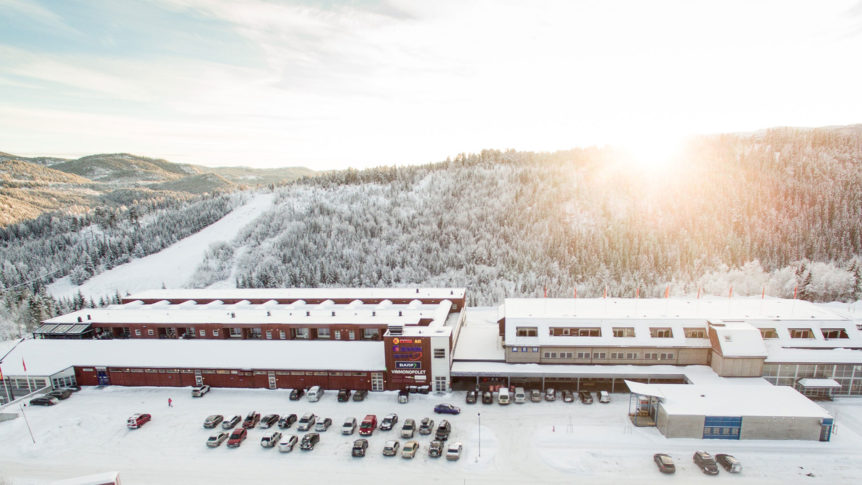 Dronefoto av snødekt bygning omringet av skog og fjell i solnedgang. Parkerte biler foran. Vinterlandskap.