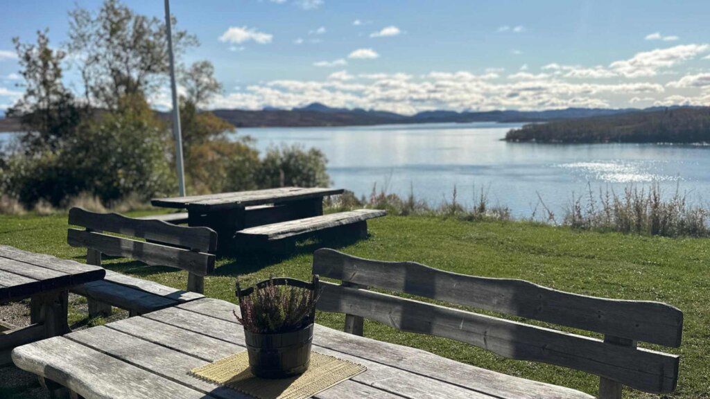 Utsikt over en innsjø med benker og bord i forgrunnen, omgitt av natur og blå himmel.