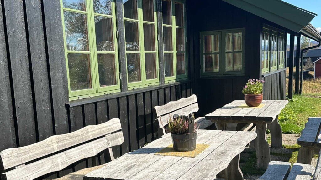 Trebord og benker foran et svart trehus med grønne vindusrammer, dekorert med blomsterpotte, solfylt dag.