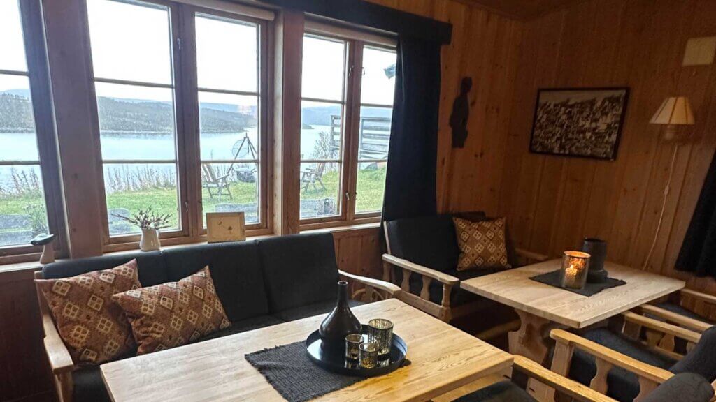 Koselig stue med treverk, komfortable sofaer og stort vindu med utsikt mot innsjøen. Stemningsfull belysning.