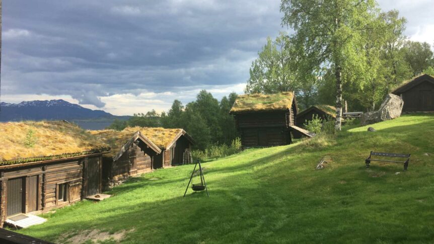 Historiske tømmerhytter med torvtak i norsk landskap, omgitt av grønn natur og fjell i bakgrunnen.