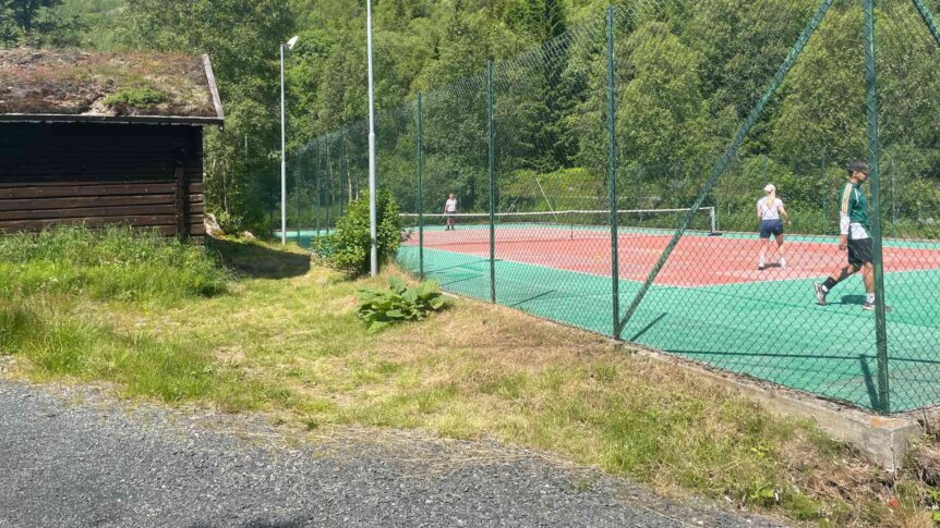 To personer som spiller tennis på utebaner, med en bygning i bakgrunnen og frodig grønt landskap.