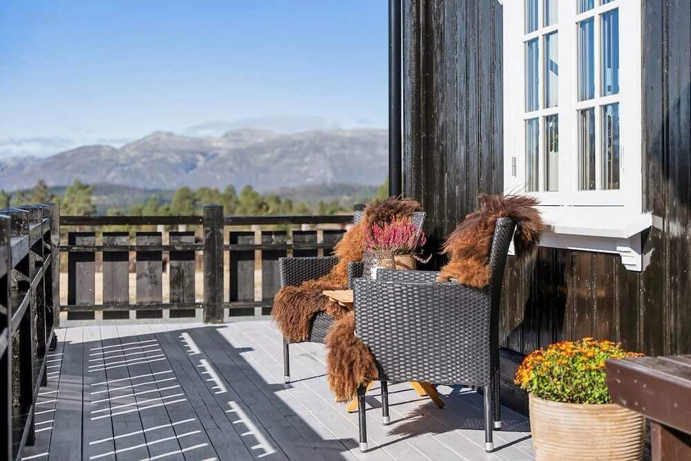 Utsikt over fjell fra terrasse med to stoler og bord, dekorert med pelser og blomster, solfylt dag.