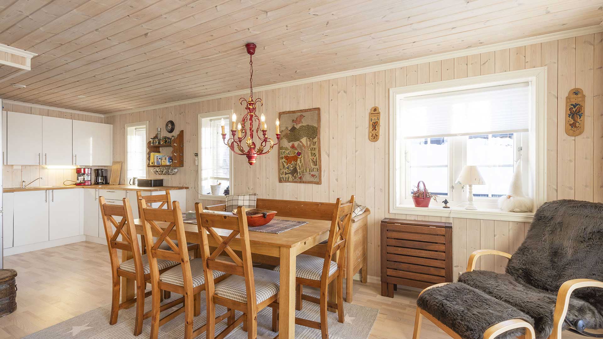 Koselig spisestue med trepanel, rødt lysekrone, og hvitt kjøkken i klassisk norsk hyttestil.