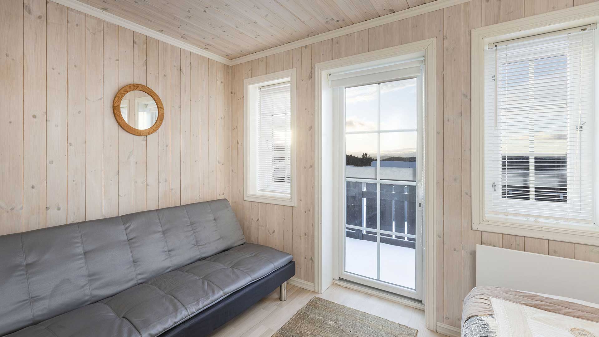 Lyst rom med grå sofa, trepaneler, speil og utsikt over vinterlandskap gjennom store vinduer.