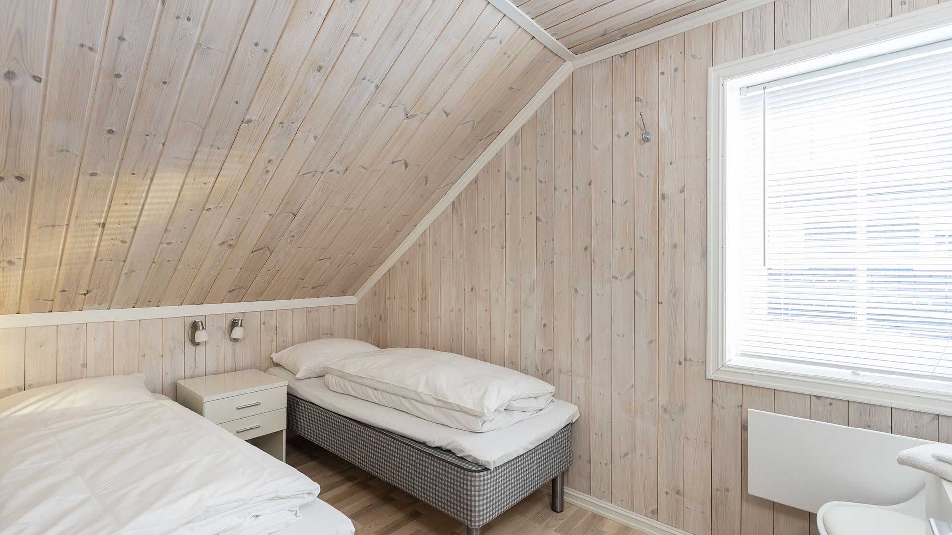 Koselig soverom med skråtak, to senger, lyse furupaneler og naturlig lys fra vinduet.