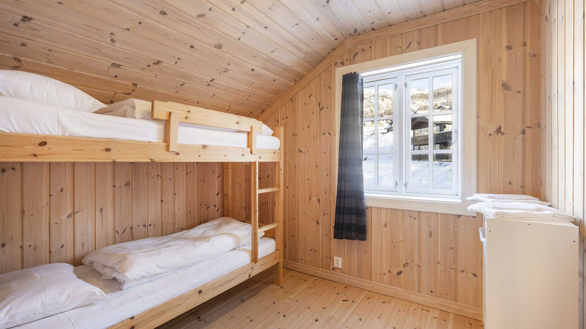 Koselig soverom med køyesenger i treverk, hvitt sengetøy og utsikt over snødekket landskap gjennom vinduet.