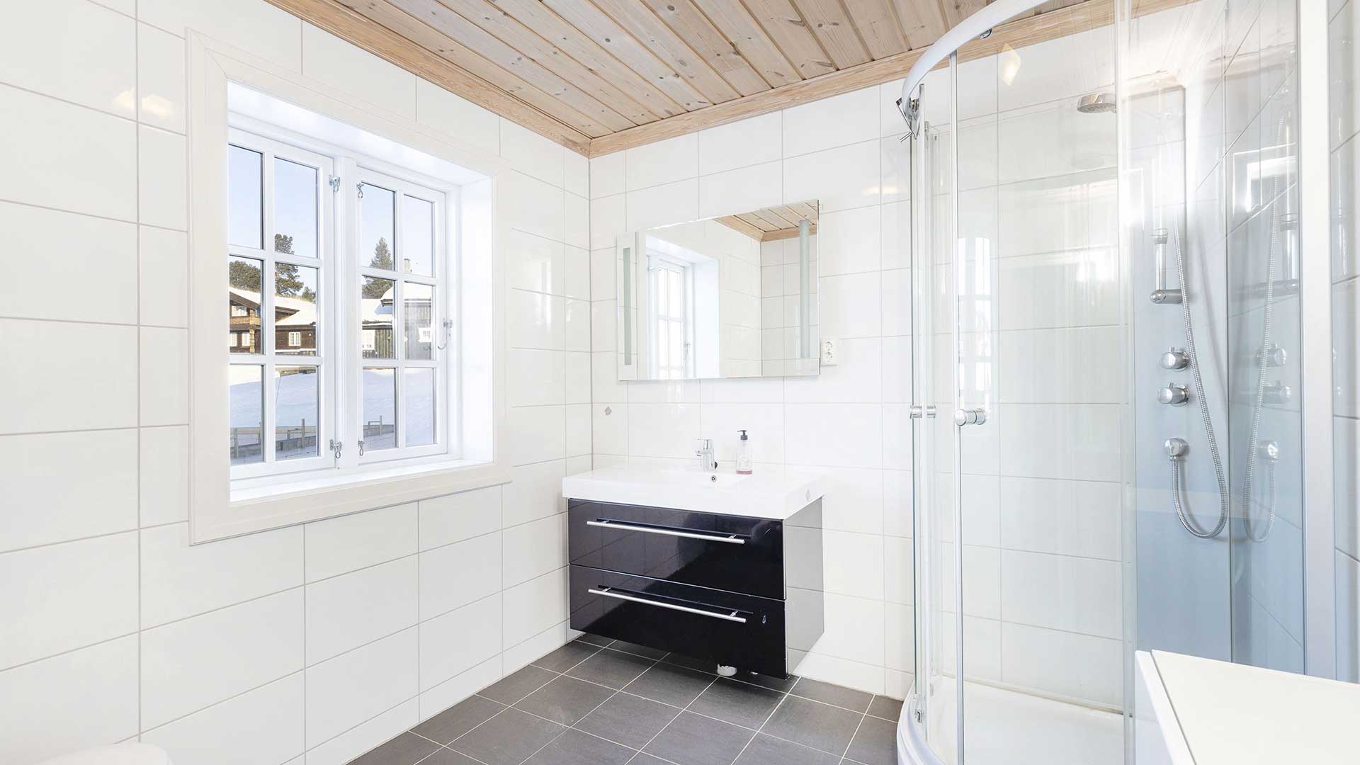 Lyst og moderne bad med dusjkabinett, vindu og svart servantskap. Fliser på vegger og gulv, trepanel i taket.