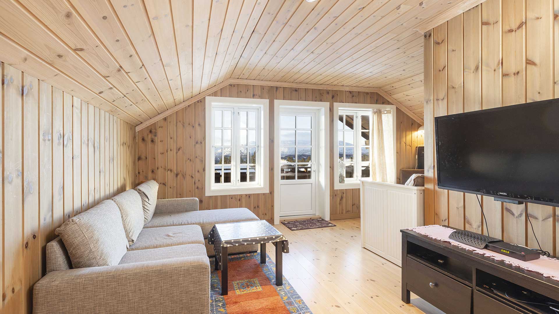 Koselig stue med trepanel, sofa, TV og store vinduer med utsikt, perfekt for avslapning i en fjellhytte.