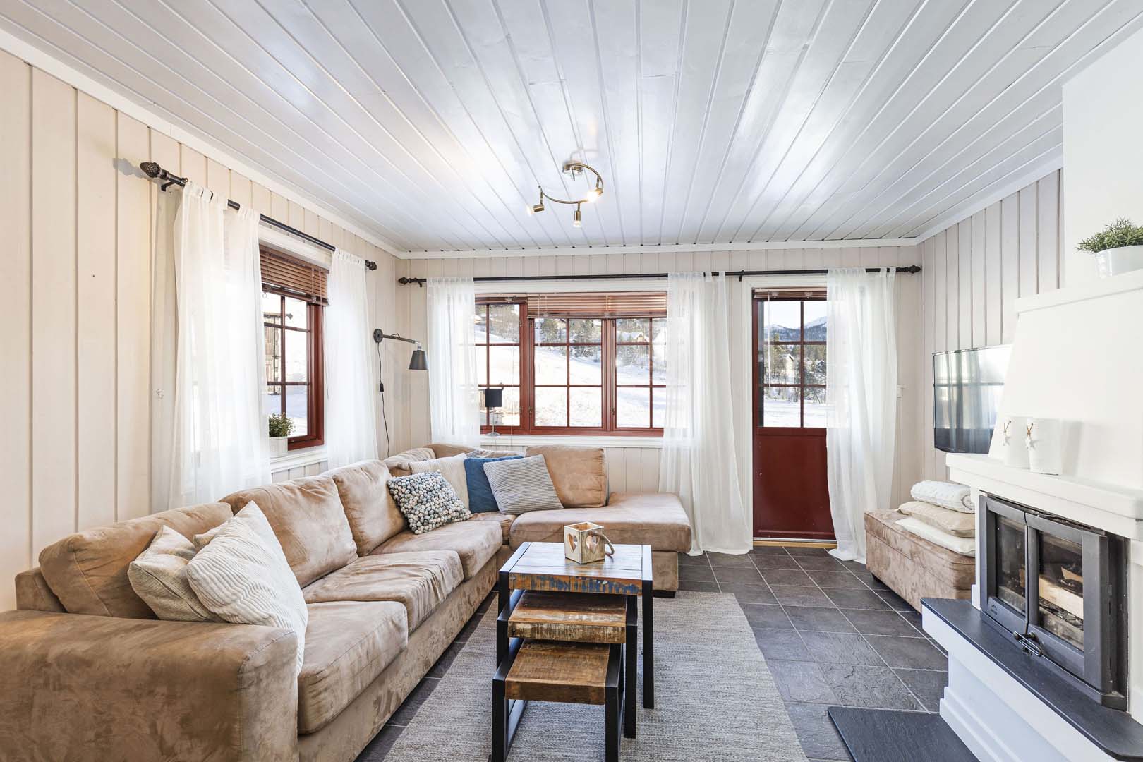 Koselig stue med beige sofa, peis og store vinduer med utsikt mot snødekte fjell. Lys innredning og naturlig lys.