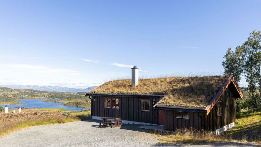 Hytte med torvtak i norsk landskap, med fjell og innsjø i bakgrunnen under blå himmel.
