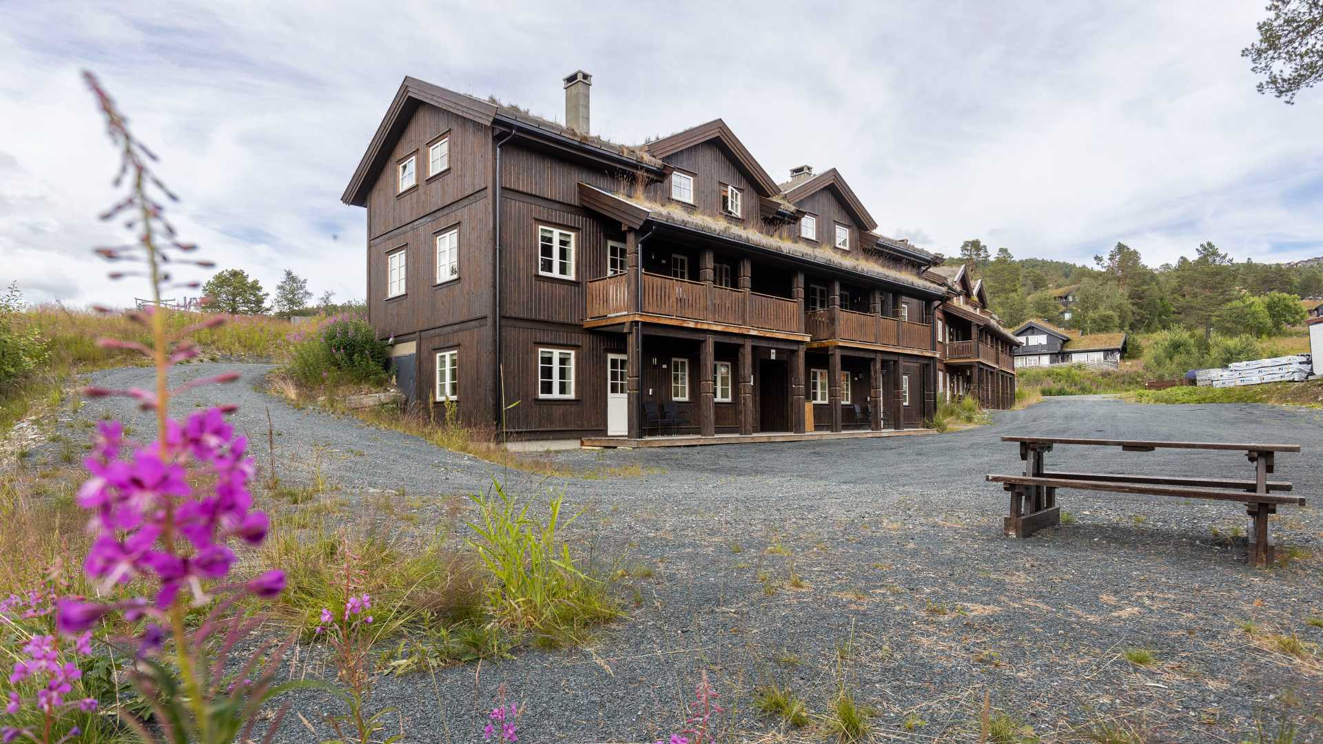 Mørk trebygning med blomster i forgrunnen, omgitt av grønt landskap og grusvei under overskyet himmel.