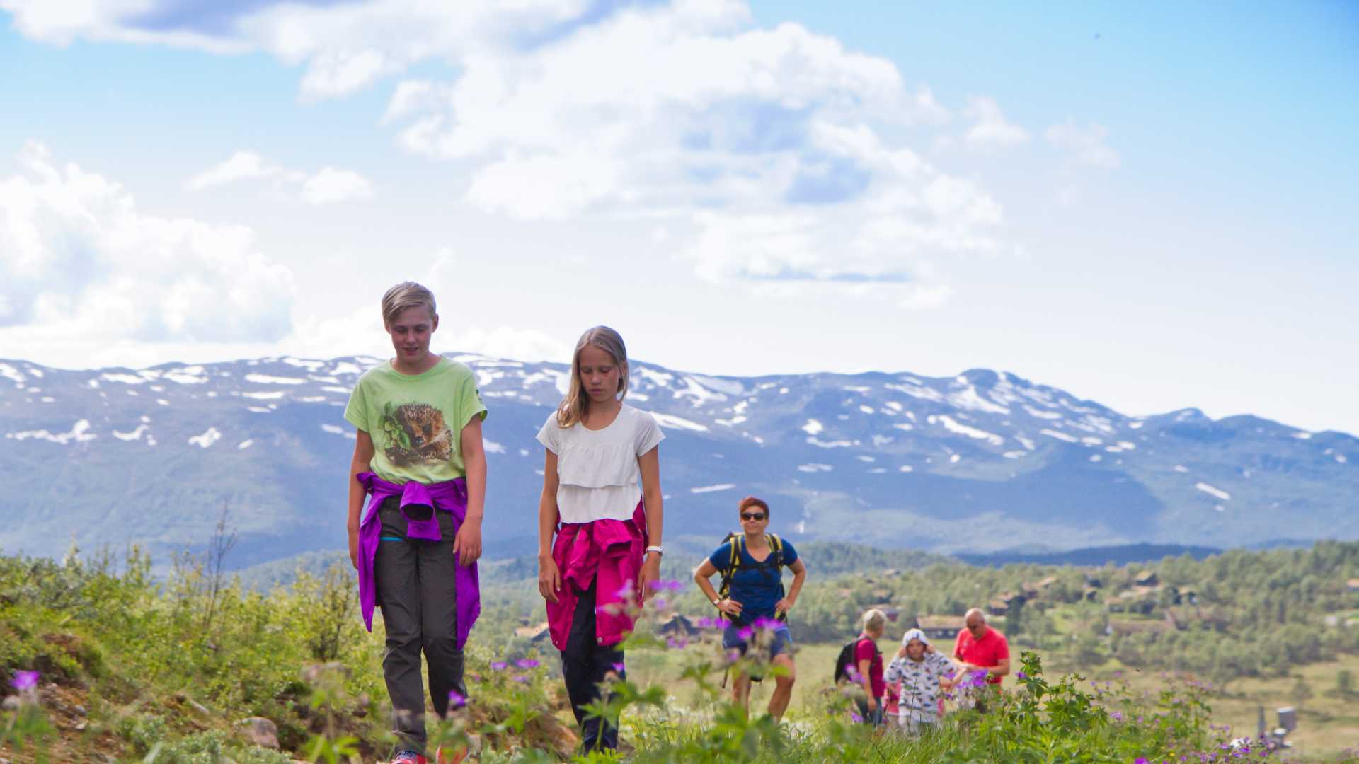 Barn og voksne på tur i norsk fjellandskap med snødekte topper og blå himmel.