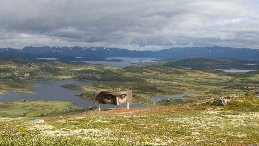 Fjellandskap med innsjøer og markant kunstverk av ansikt plassert i midten, under en dramatisk himmel.