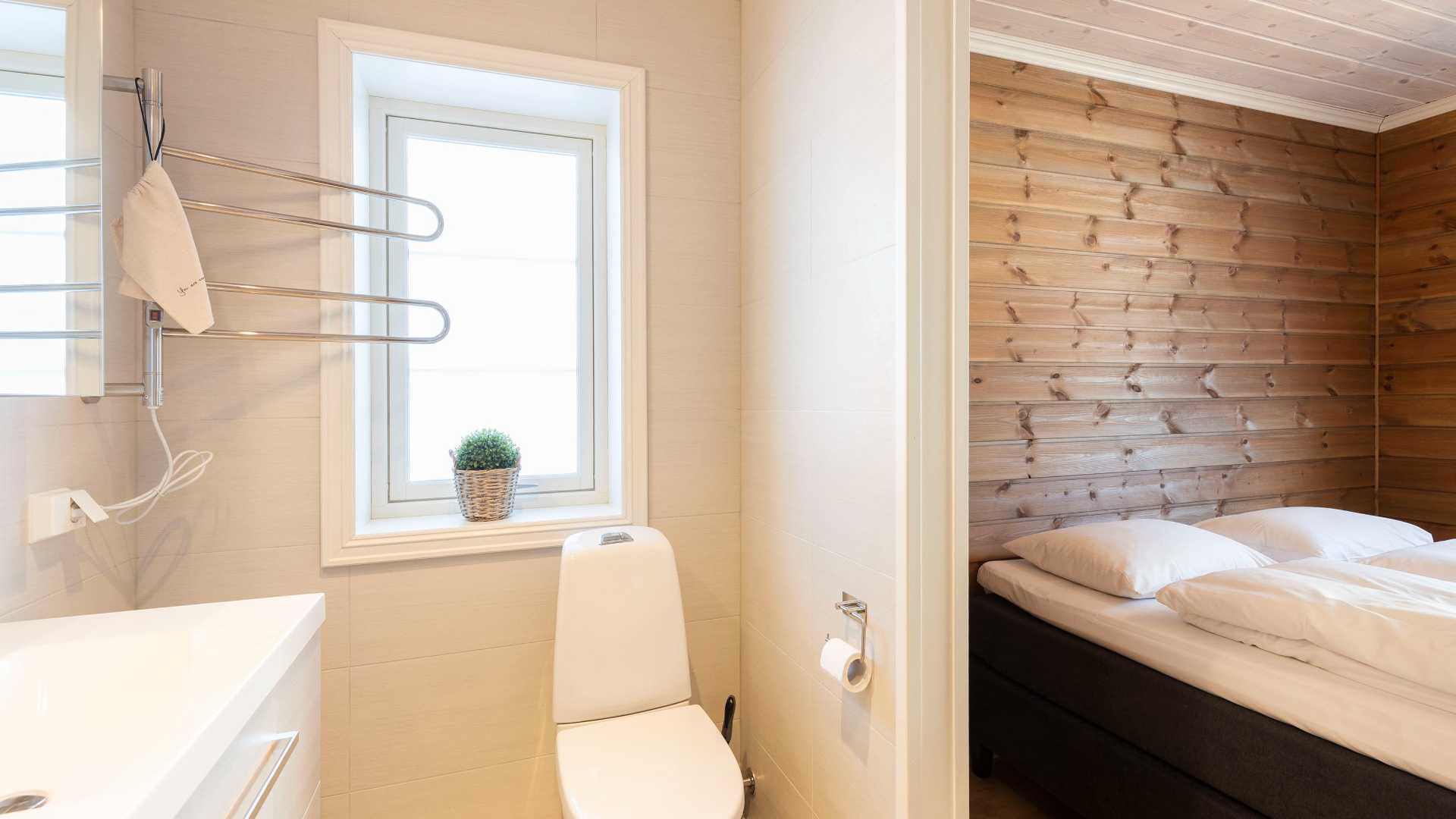 Lyst bad og soverom med trepanel, toalett, servant, håndklestativ og planter ved et vindu. Komfortabel og moderne innredning.