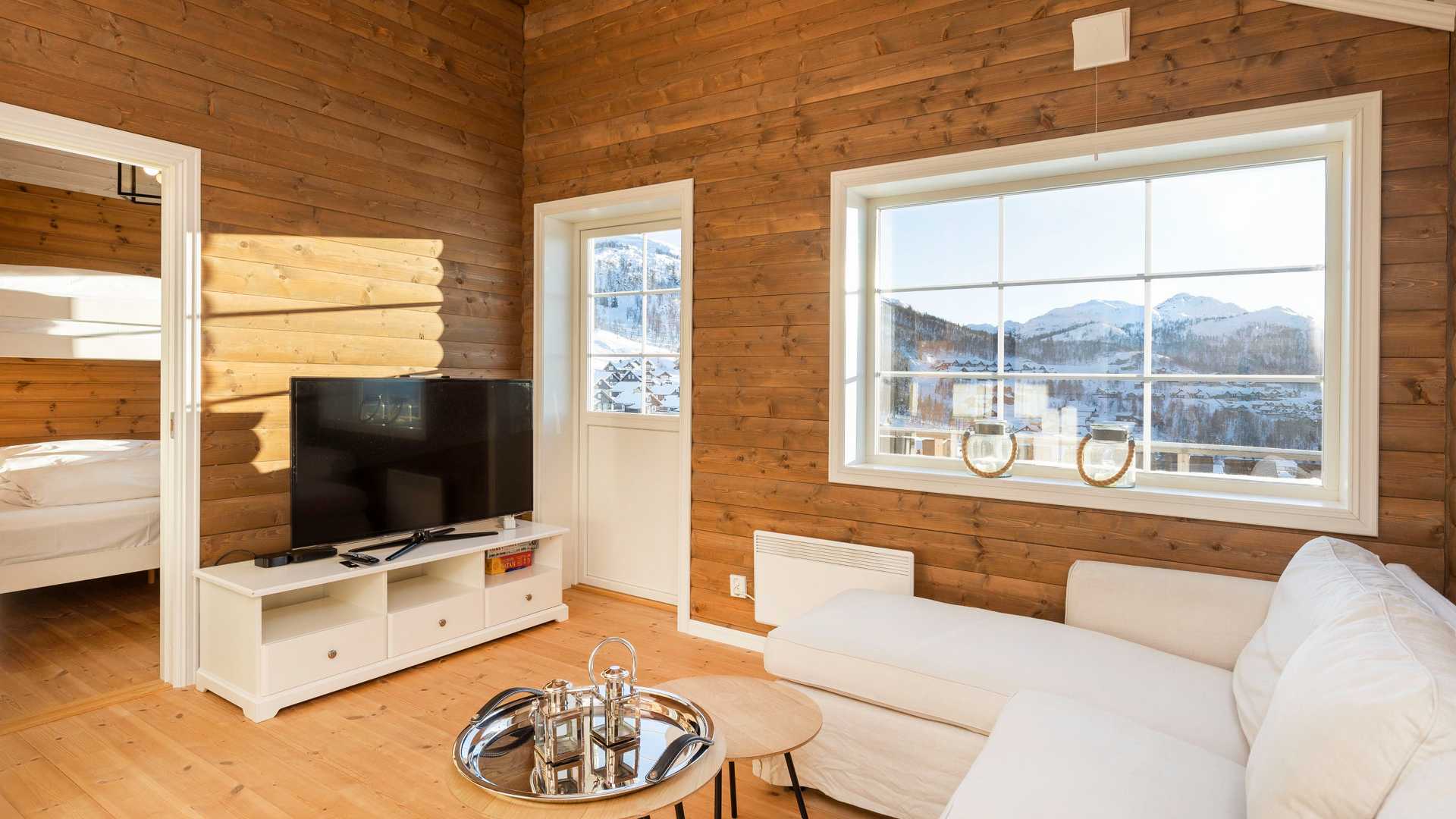Moderne stue med trepanelvegger, stor TV og fjellutsikt gjennom vinduet, ved siden av et komfortabelt, hvitt hjørnesofa.