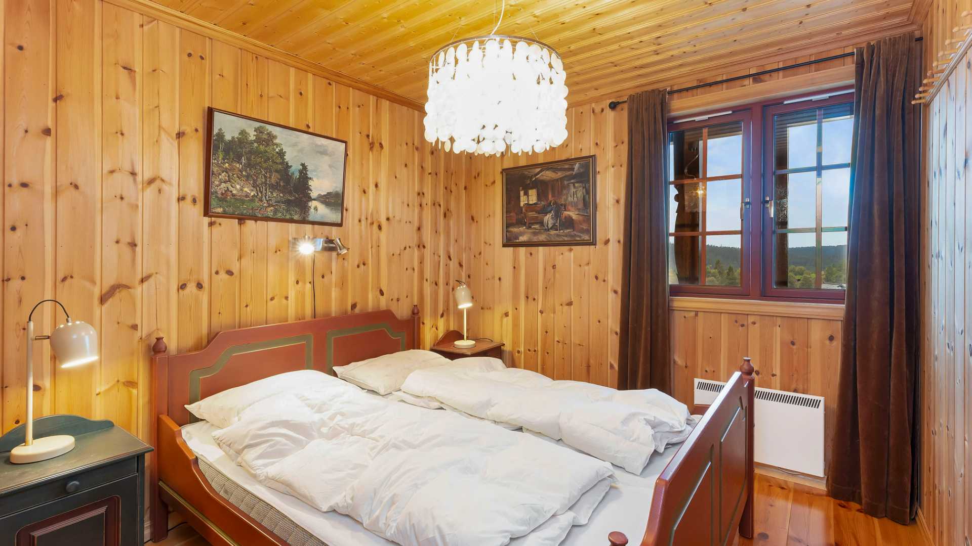 Koselig soverom med trepaneler, dobbeltseng, moderne lysekrone og vindu med utsikt mot naturen.