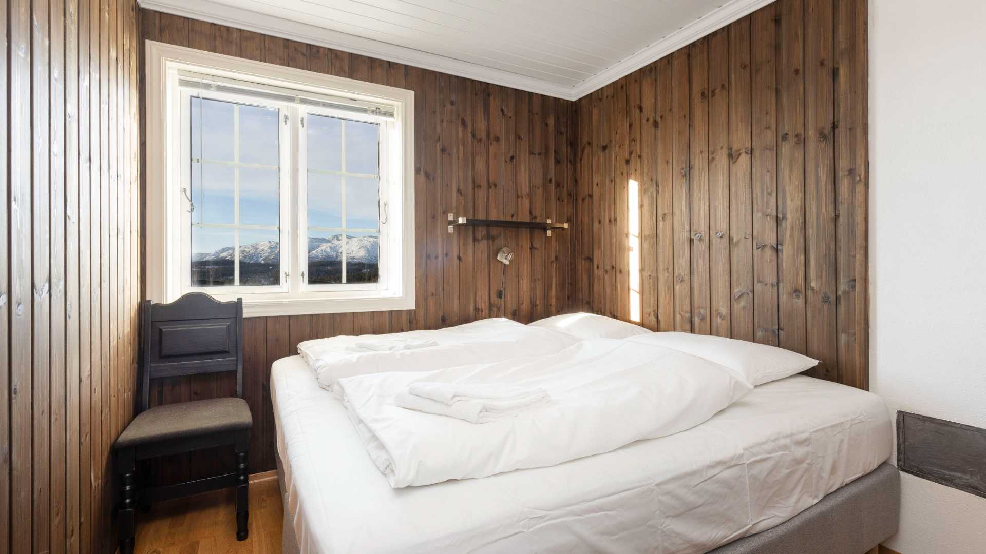 Lyst soverom med trepanel, dobbeltseng og utsikt mot snødekte fjell gjennom vindu. Rustikk hytteinnredning.