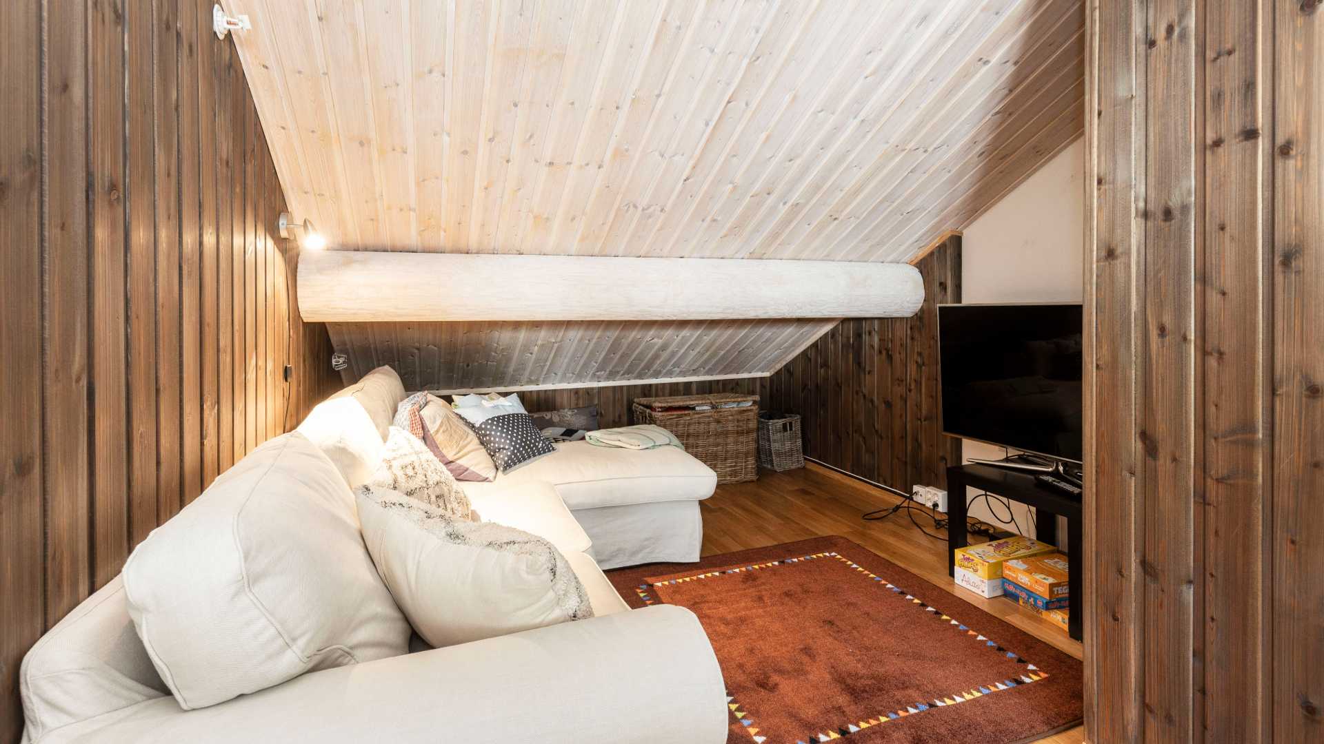 Koselig loftstue med skråtak, beige sofa, TV og trevegger, perfekt for avslapning.