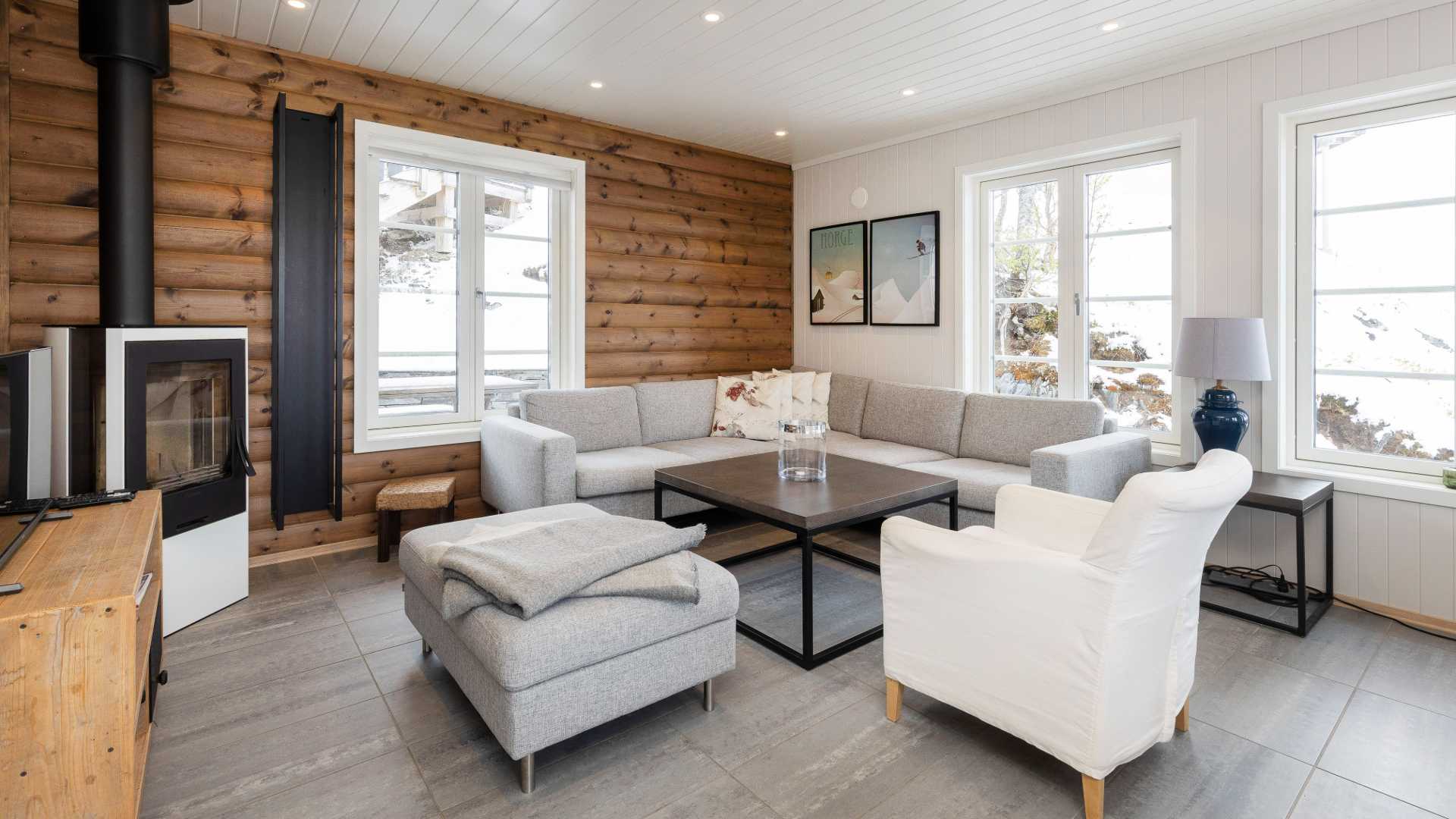Moderne stue med grå sofa, peis og store vinduer som gir utsikt til snødekt landskap. Koselig hytteatmosfære.