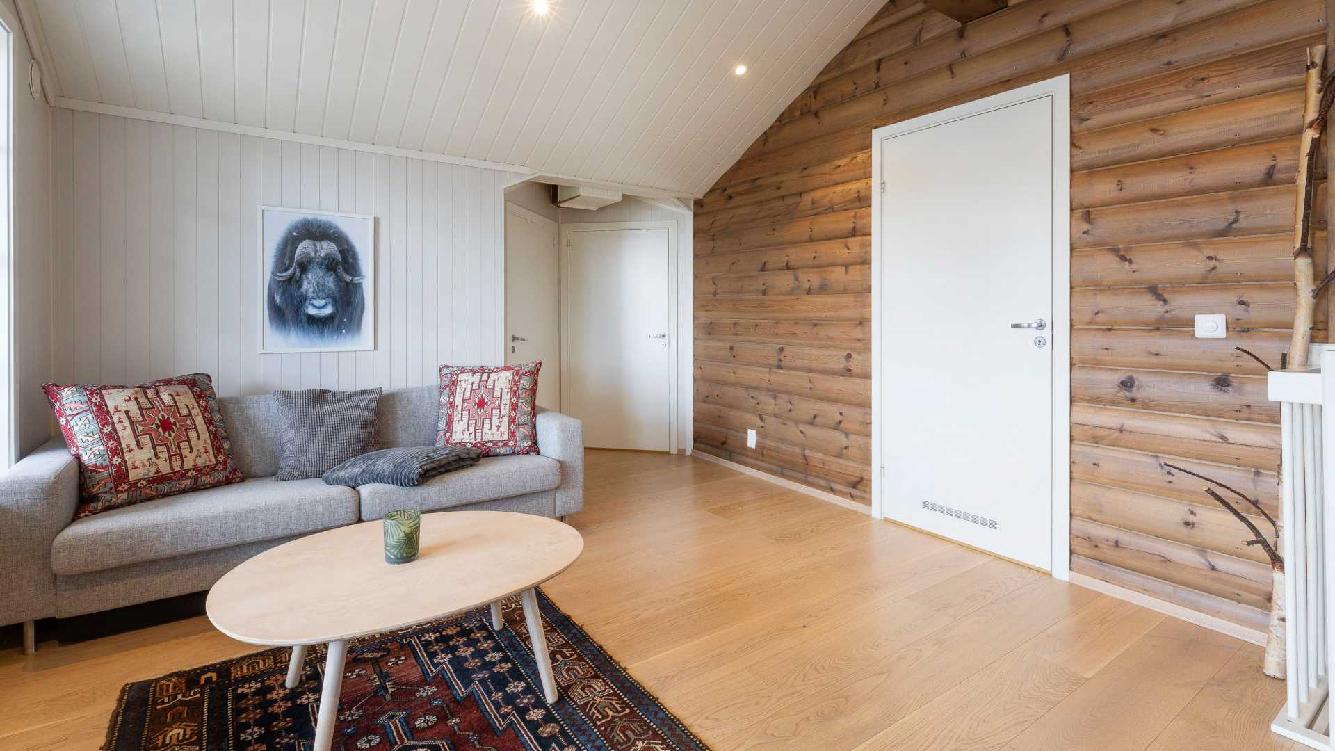 Stue med grå sofa, tregulv og trepanelvegger, dekorert med puter og et dyrepresentert bilde på veggen.
