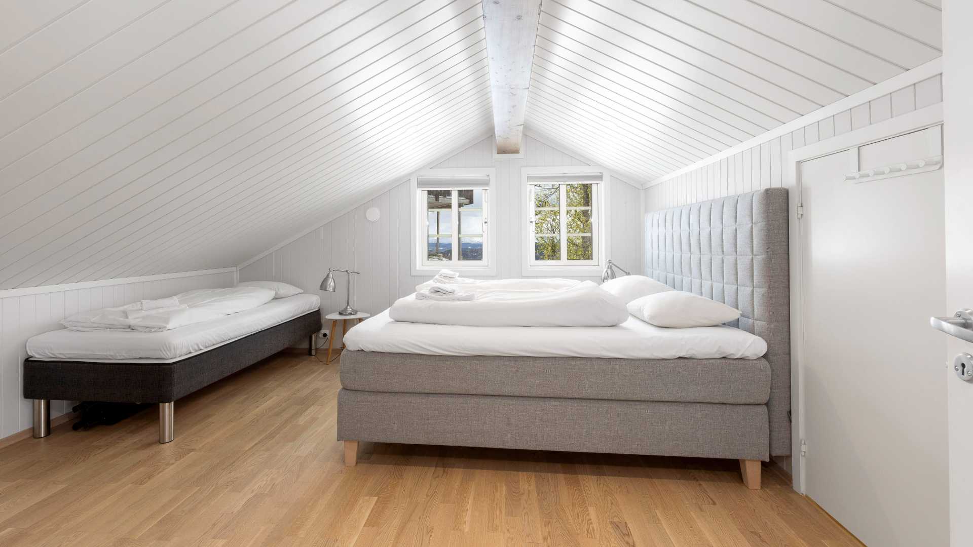 Lyst soverom med skråtak, to senger og utsikt ut vinduet. Elegant design med hvite paneler og tregulv.