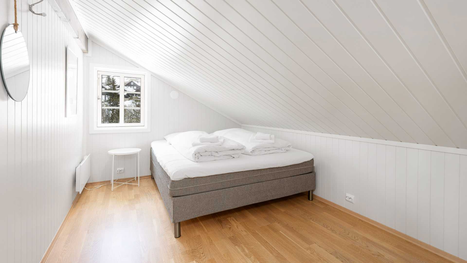 Lyst og koselig soverom på loftet med dobbeltseng, hvite vegger og tregulv.